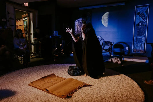 image from moon witch 2022. a woman in a cloak stands on stage, lit in blue light, arms raised, with a moon projected behind her and an oversized book on the floor