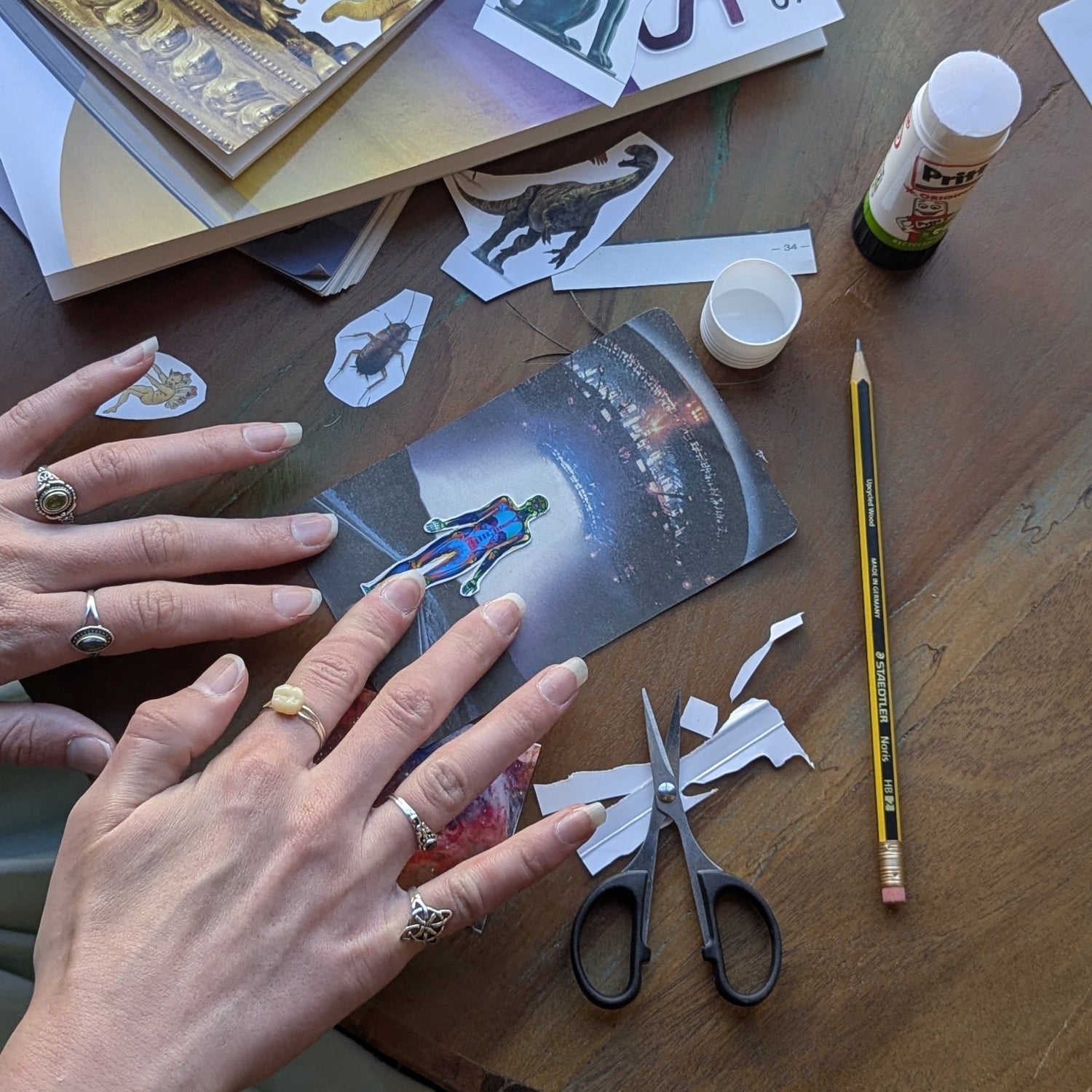 A top-down view of a wooden desk with hands working on a craft project, cutting a printed card with scissors and tape. The desk is cluttered with books, stickers, a pencil, and a small bottle of glue.