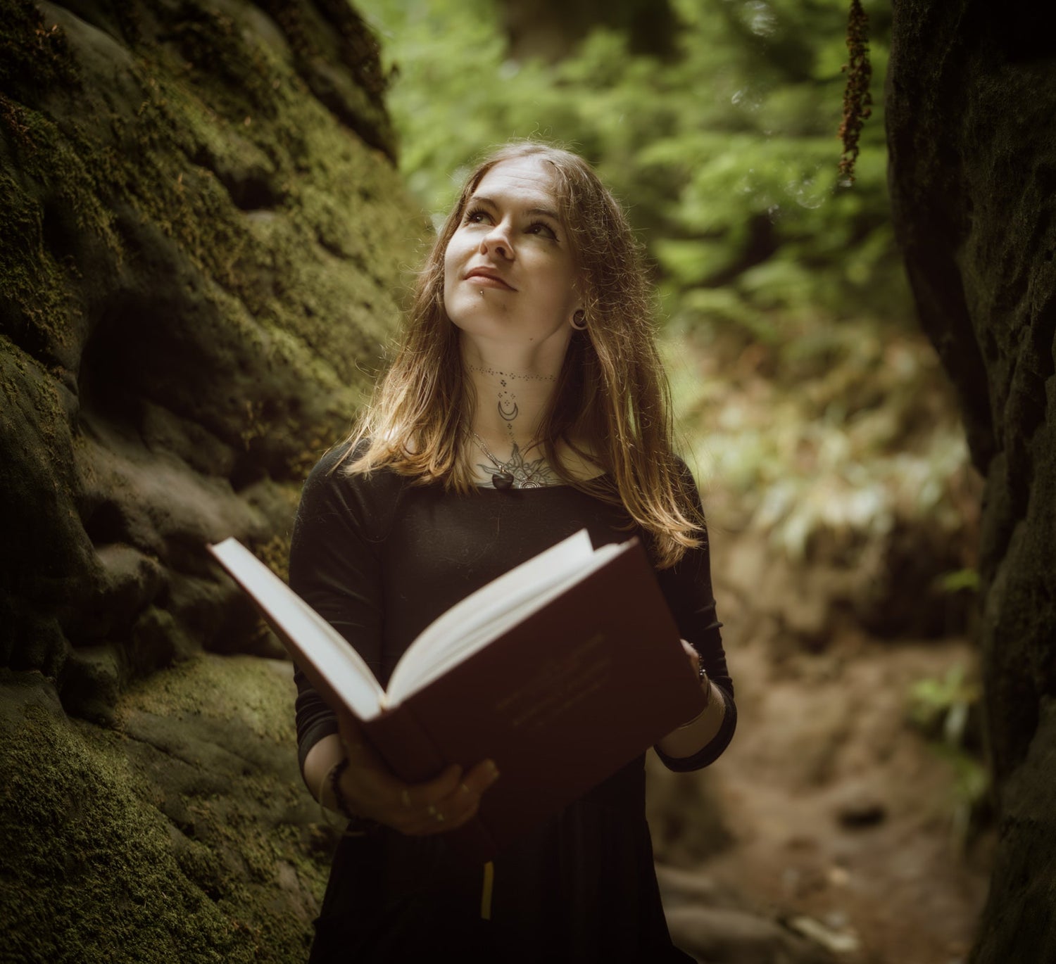 A woman with long light brown hair stands in a mossy stone passage holding an open book, looking upward in a forest setting.