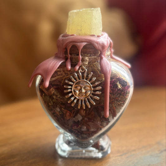 A small clear glass heart-shaped bottle filled with dried botanicals, topped with dripping pink wax and a pale yellow crystal plug, sitting on a wooden surface.